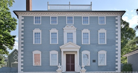 Image Of A Three Story Blue House And White Trimmed Windows With A White Fence In The Foreground.